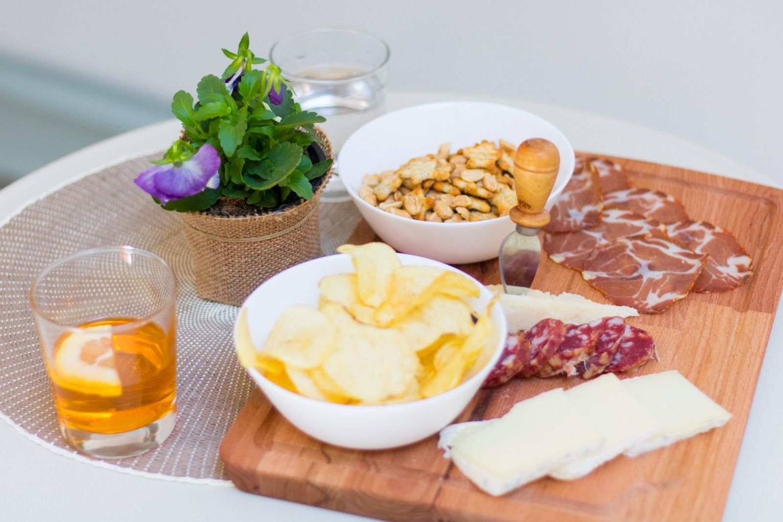 A wooden board with various food items including cheese, cured meats, nuts, and chips, accompanied by a glass of amber liquid and a small potted plant.