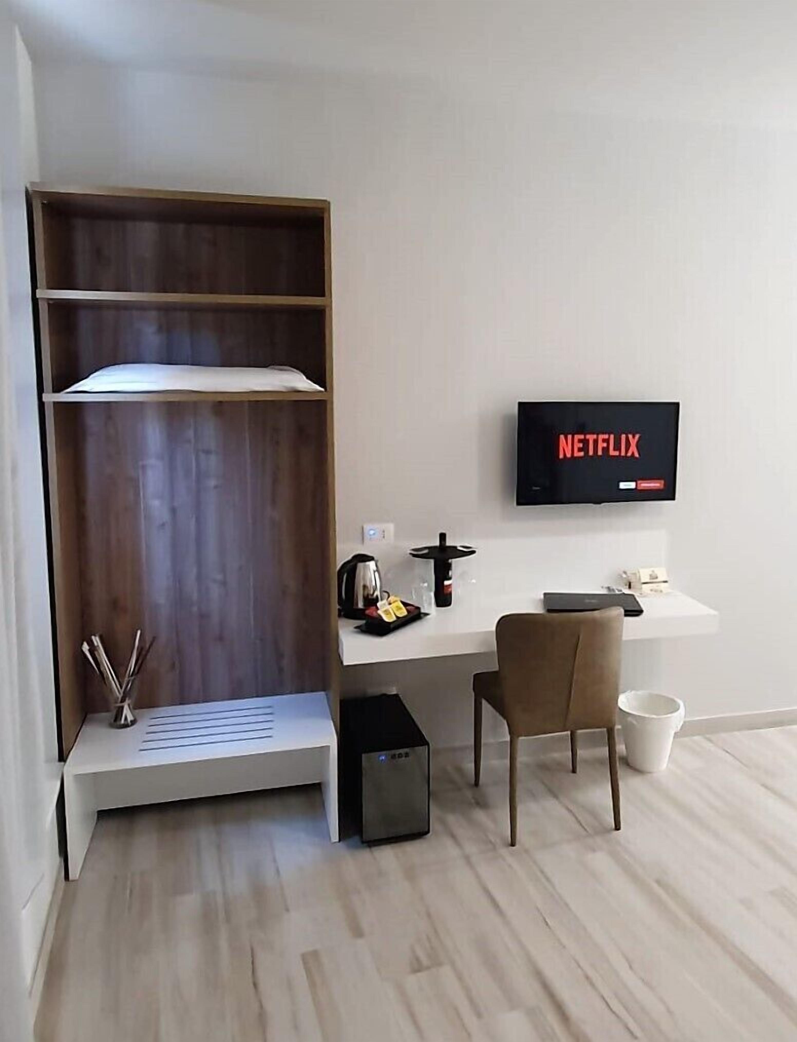 Una stanza con un'unità a scaffali in legno, una scrivania con una sedia, un televisore che mostra 'NETFLIX' e una macchina per il caffè.