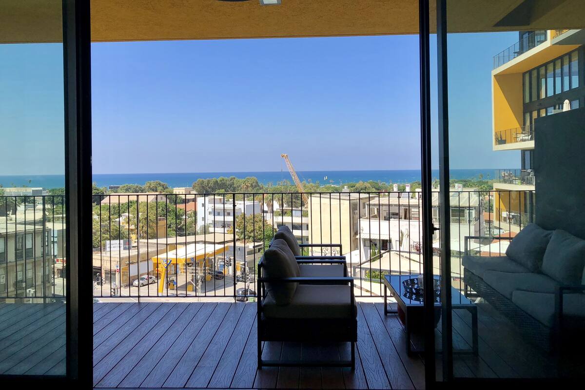 A balcony with a view of the sea and construction cranes, a sofa, and a table.