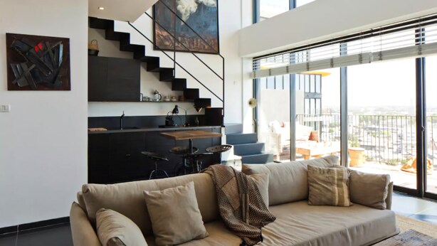 A modern living room with a sofa, a staircase leading to a kitchen, and large windows offering a view of the city. - Elifelet 26 (Tel Aviv)