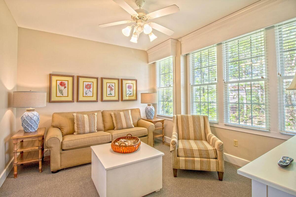 A living room with a beige sofa, striped armchair, and a coffee table. There are framed pictures on the wall, a ceiling fan, and large windows with blinds.