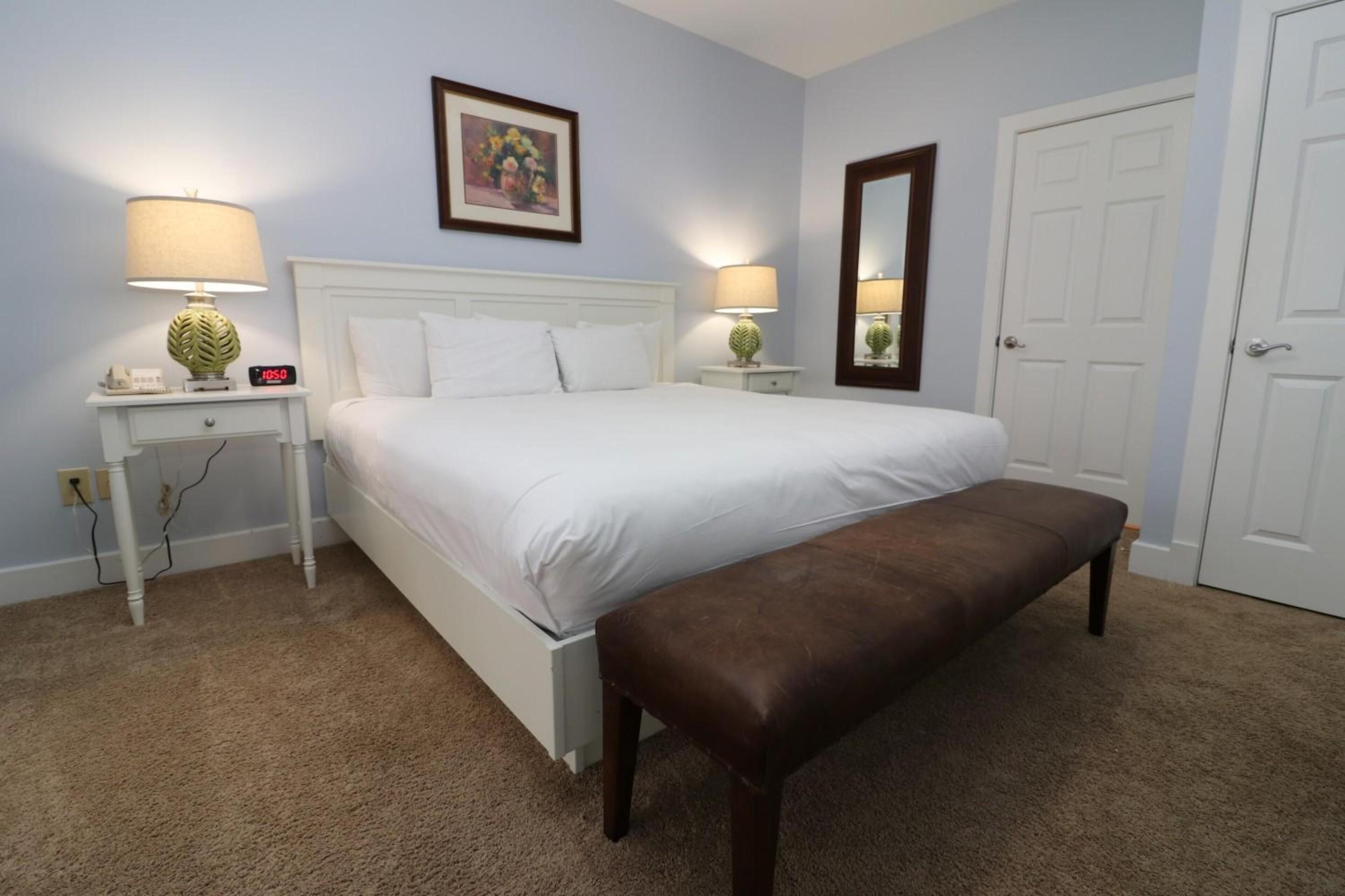 A bedroom with a bed, two bedside tables with lamps, a bench, a door, and a wall-mounted picture.