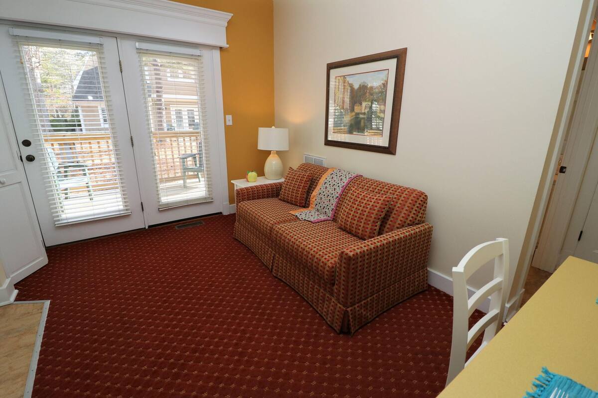 A room with a patterned red carpet, a brown checkered sofa, a white chair, a yellow table, and a framed picture on the wall.