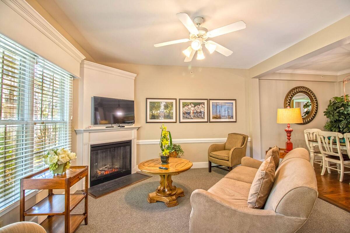 A living room with a fireplace, a ceiling fan, a dining area with chairs, and a television.