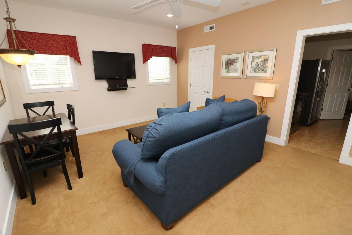 A living room with a blue sofa, a TV, a dining table with chairs, and a lamp.