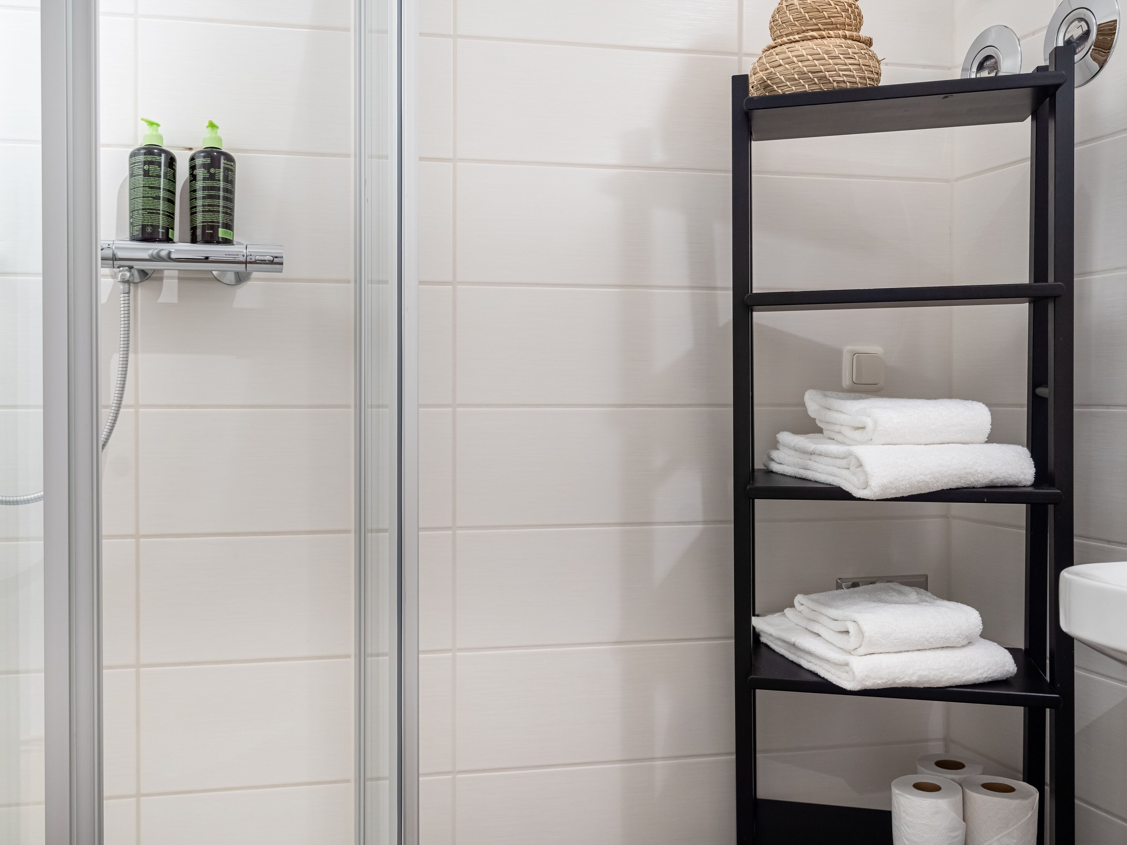 A bathroom with a glass shower enclosure, a black shelf with toiletries, and a black shelf with towels and toilet paper.