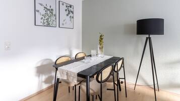 A dining area with a table set for four, surrounded by wicker chairs. There are two framed botanical prints on the wall, a vase with a single plant on the table, and a floor lamp to the right.