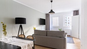 A modern living room with a grey sofa, a dining table with chairs, a floor lamp, and a TV mounted on the wall.