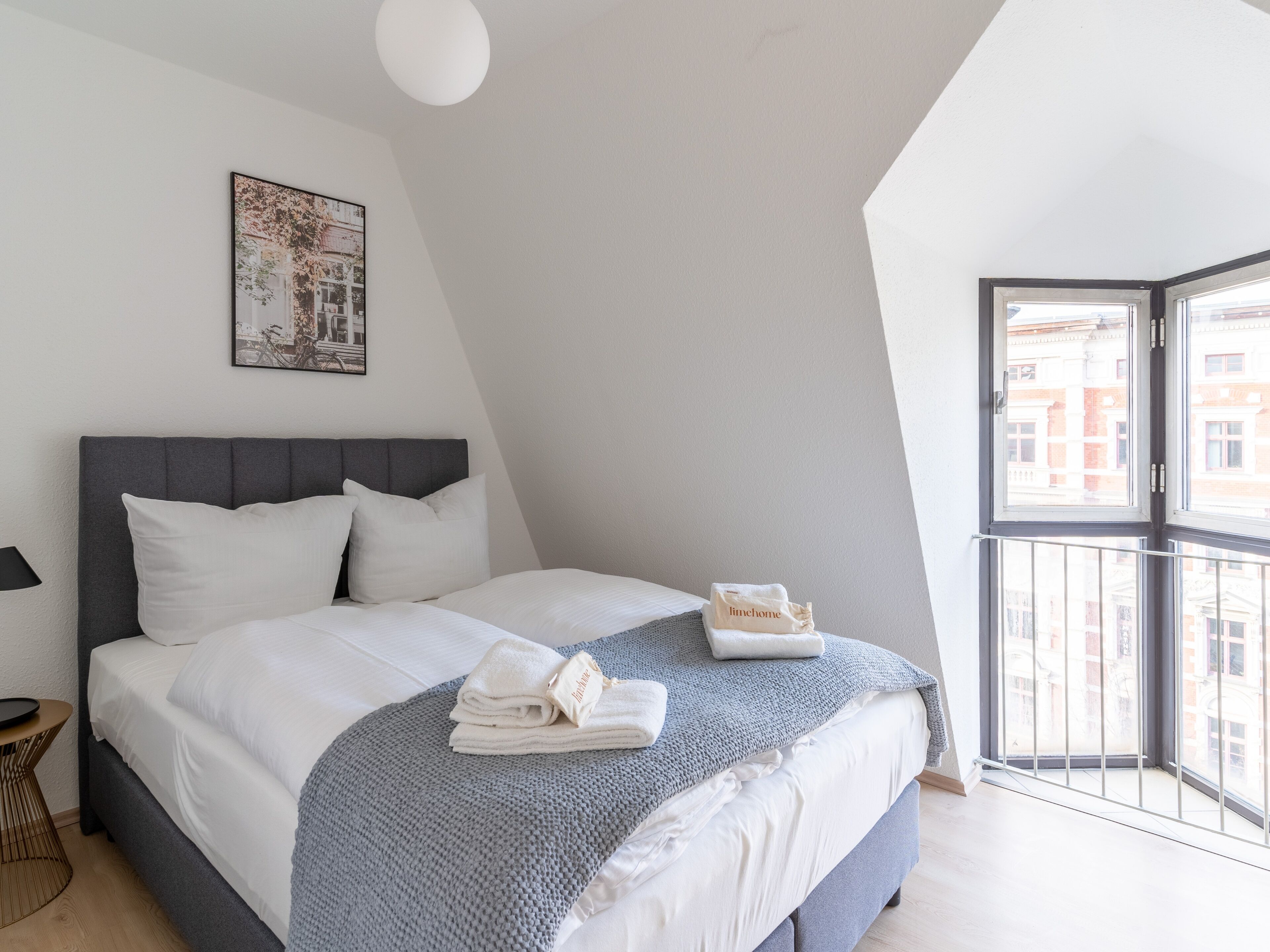 A modern bedroom with a grey headboard, white bedding, a grey knitted throw, and a view of buildings through a large window.