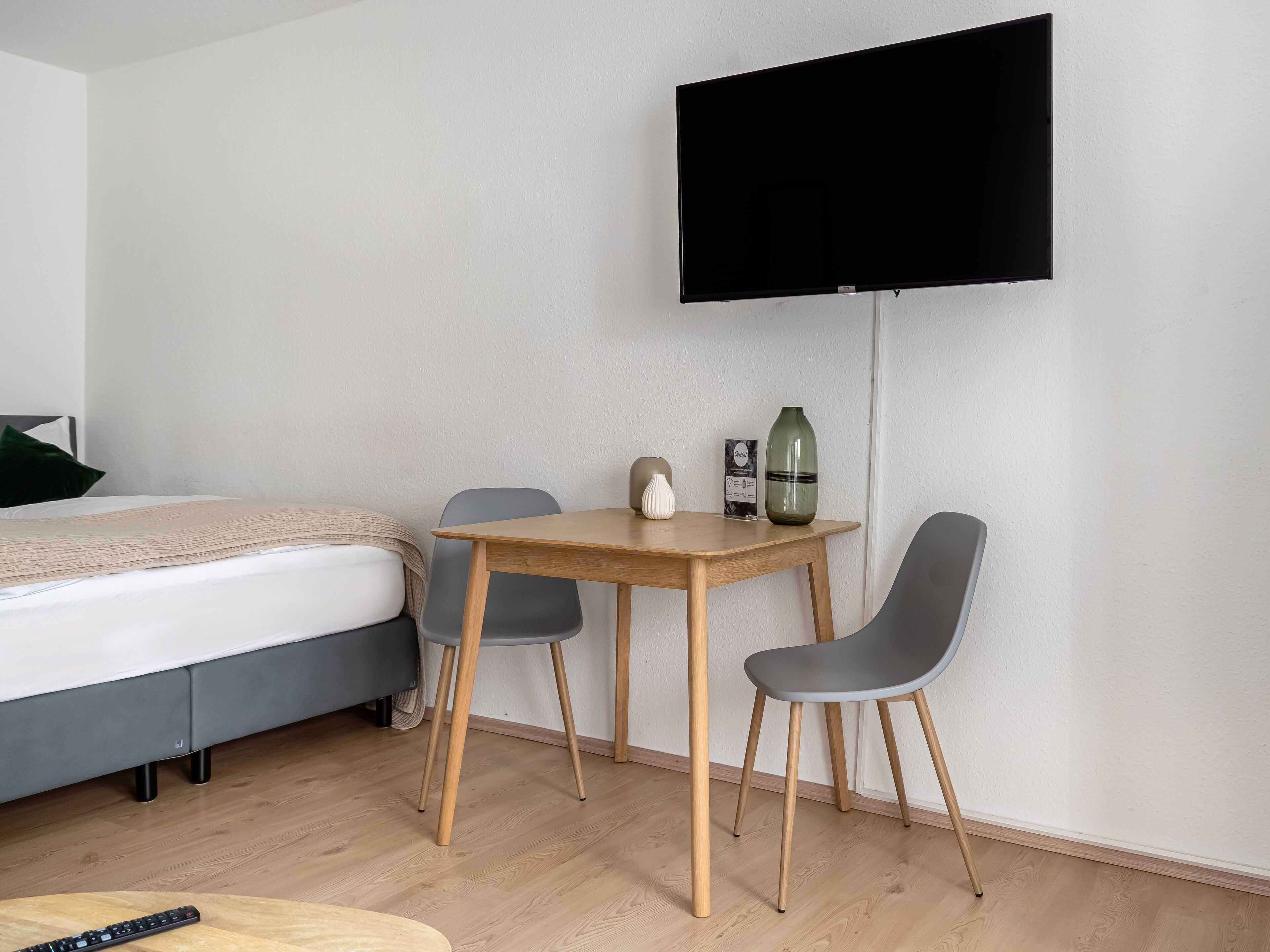 A hotel room with a bed, a wooden table with two chairs, a TV mounted on the wall, and a vase on the table.