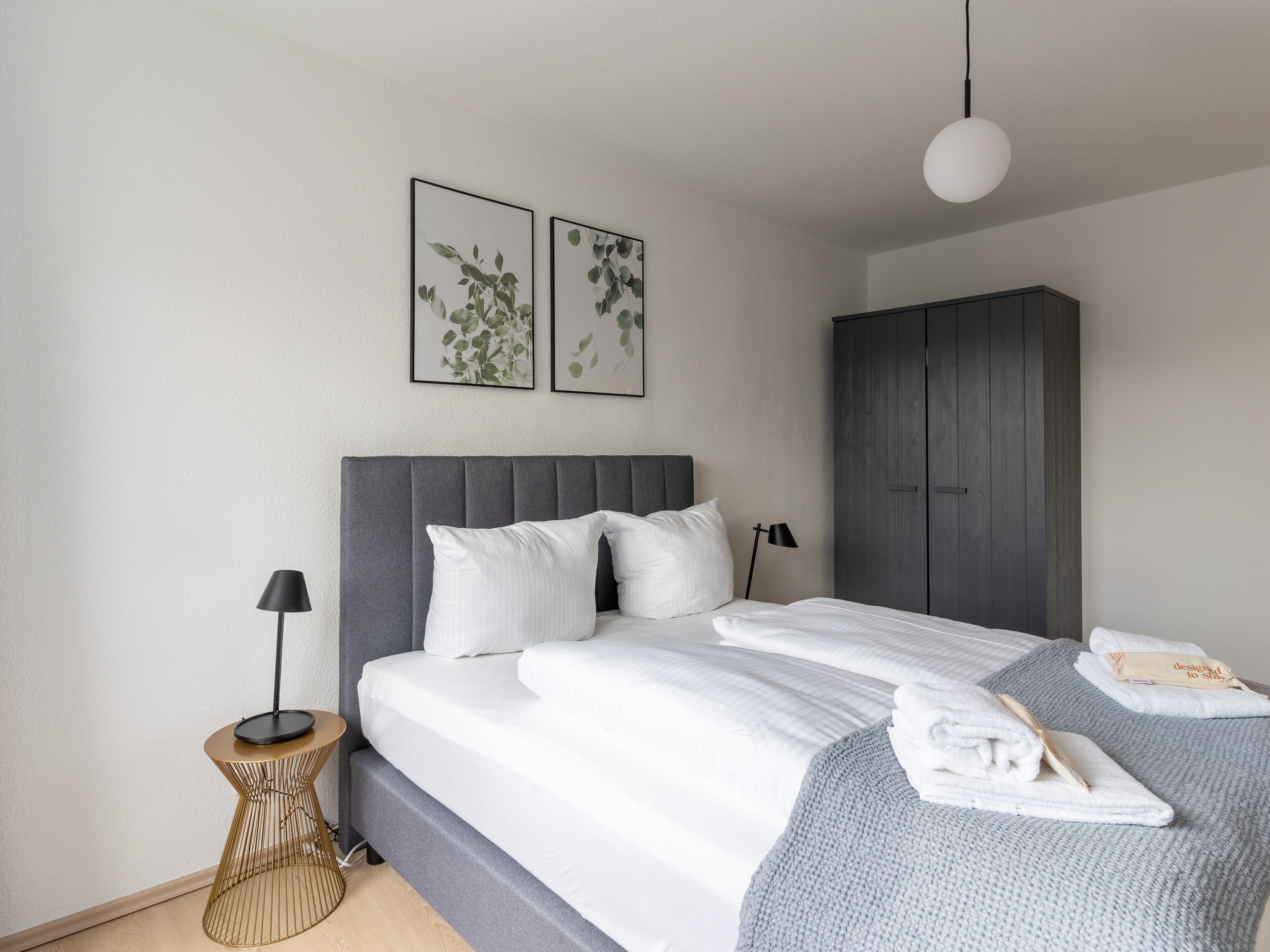 A modern bedroom with a grey upholstered headboard, a round bedside table with a lamp, a bed with white linens, and a grey knitted throw blanket.
