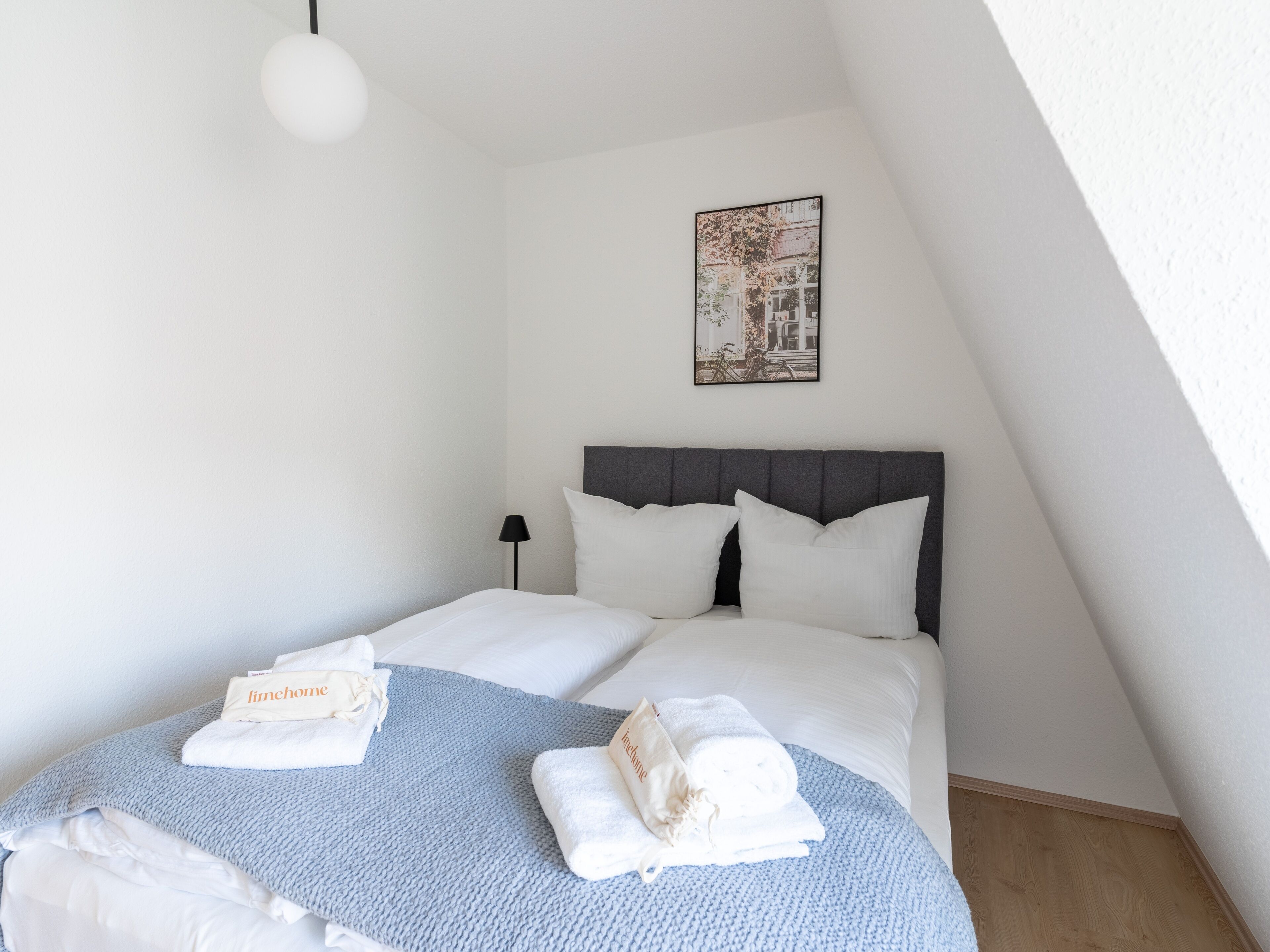 Una cama bien hecha con ropa de cama blanca y una manta azul, una lámpara de noche y un cuadro en la pared.