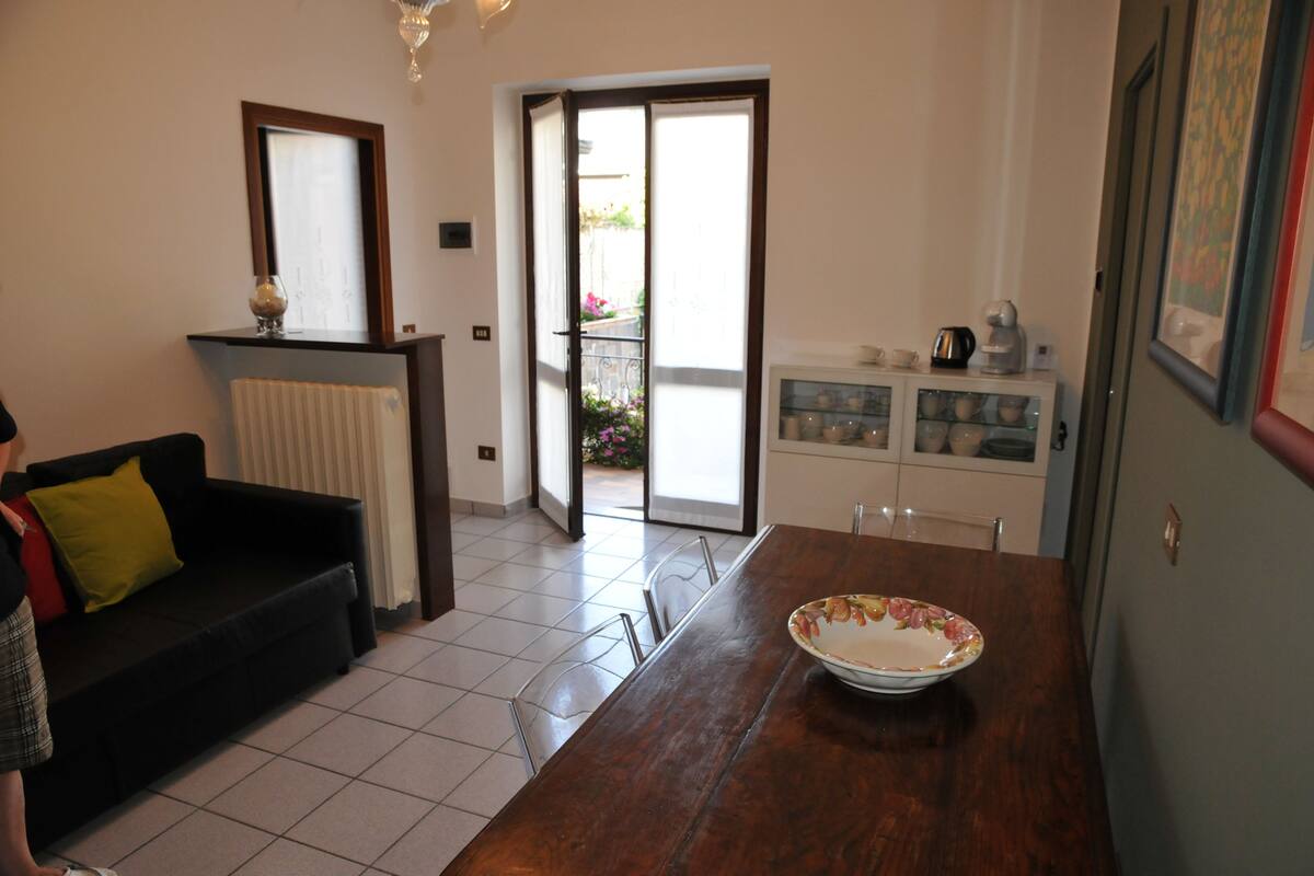 A living room with a black sofa, a wooden dining table with a bowl, a sliding glass door to a balcony, and a kitchen area with cabinets and appliances.