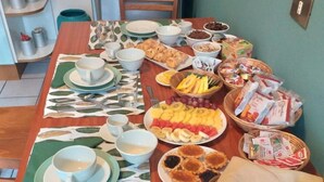 Una mesa de comedor servida con diversos platos, entre ellos frutas, pasteles y carnes, en una habitación con suelo de madera y pared verde.