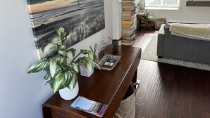 Una sala de estar con un cuadro grande de un atardecer, una mesa de centro de madera con una planta, una pila de libros y un sofá gris.