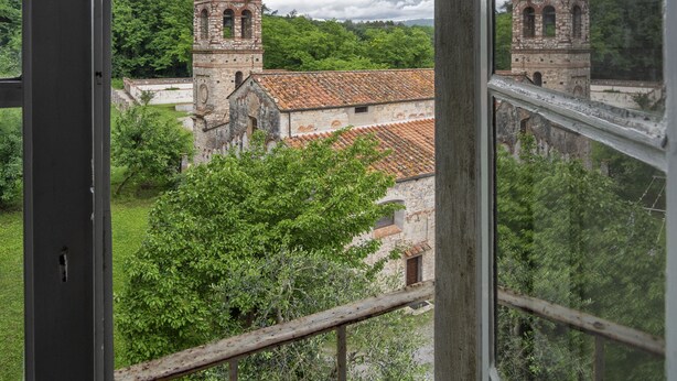 Vista di un edificio storico con due torri e un tetto in tegole rosse, ripresa attraverso una finestra con cornice in legno.