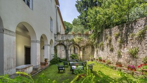 Un cortile con un edificio bianco, portali ad arco e un muro in pietra. Sono presenti piante in vaso e mobili da esterno.