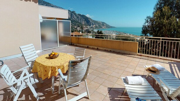 A balcony with a table and chairs, a view of the sea, and a mountain in the background. - Cozy 2BR with terrace, pool, and garage (Menton)