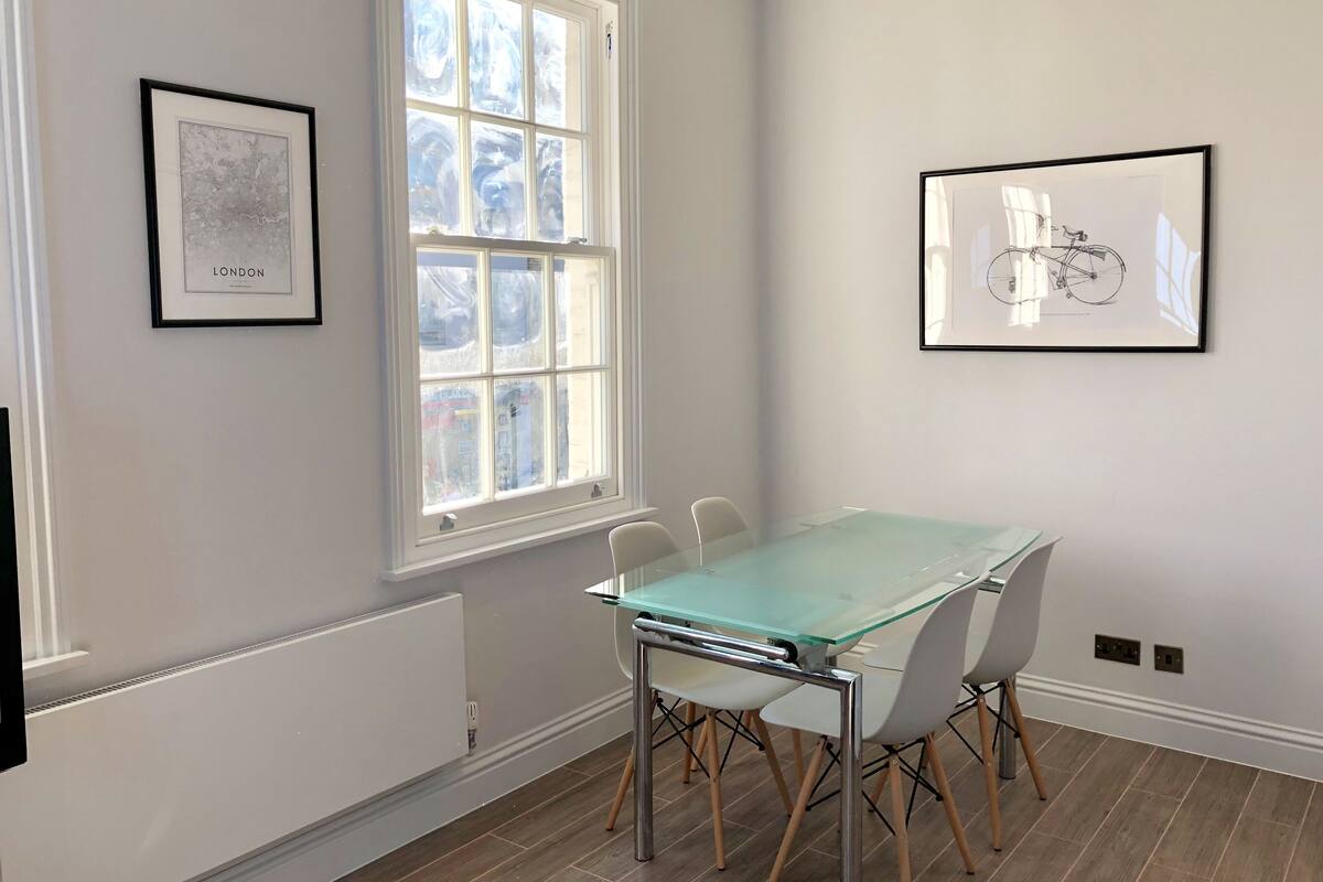 A room with a glass-top table, white chairs, a large window, and two framed artworks on the walls.