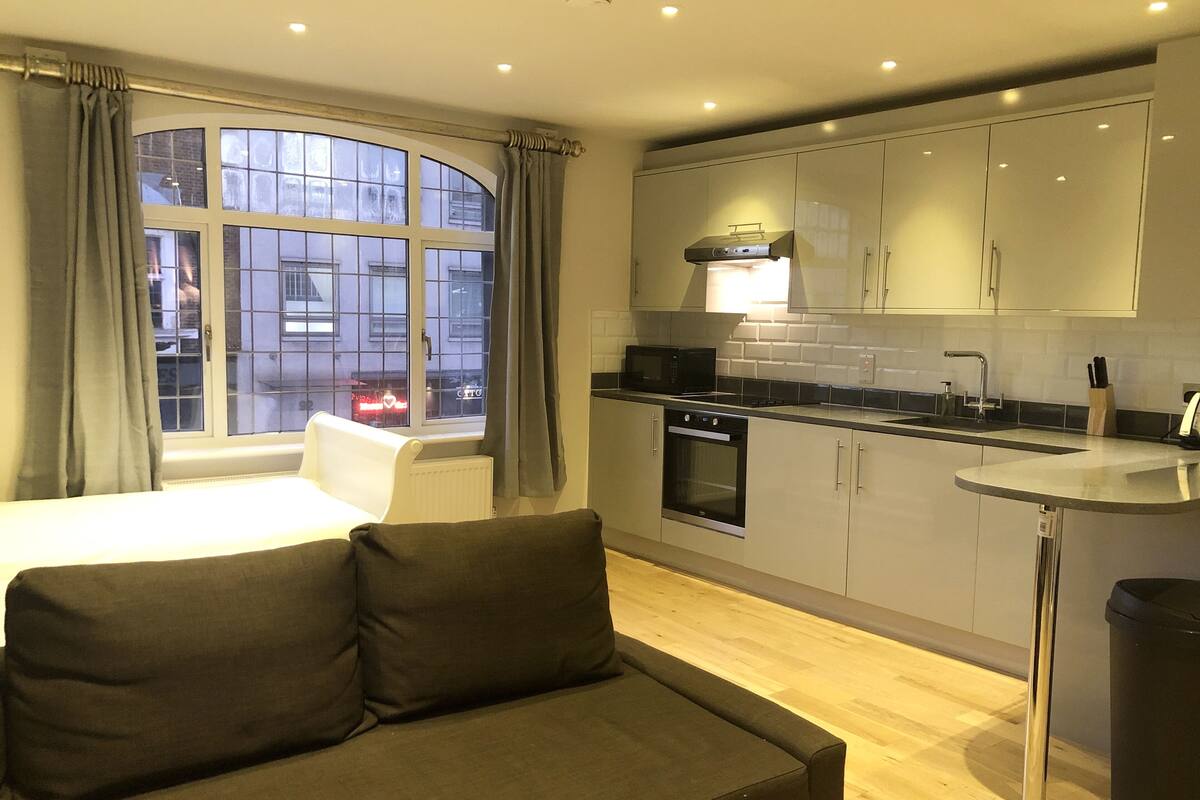 A modern kitchen with a microwave, oven, and sink. There is a window with curtains, a sofa, and a trash bin.