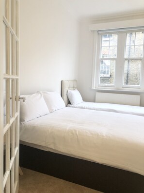 Une chambre avec deux lits, du linge de lit blanc, une fenêtre donnant sur des immeubles et une porte blanche avec des panneaux de verre.