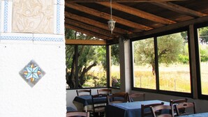 A covered outdoor dining area with wooden beams, a hanging light fixture, and a view of trees and grass through large windows. - Sweet Home La Conturella (Ricadi)