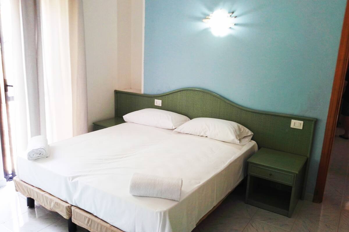 A neatly made bed with white linens, a green headboard, and a small bedside table with a lamp.