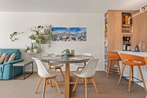 A modern living room with a round dining table, white chairs, a teal sofa, a bookshelf, and a painting of a snowy mountain landscape. - Chic 2 bed / 2 bath Apartment with City Views (Teneriffe)