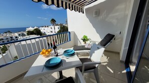A balcony with a table set for two, a chair, a potted plant, and a view of the sea and buildings.