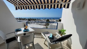 A balcony with a table set for two, a chair, a small plant, and a view of the sea and buildings. - Beautiful apartment with stunning views and communal pool (Puerto del Carmen)