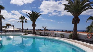 A pool with clear blue water, surrounded by palm trees and a view of the sea. - Beautiful apartment with stunning views and communal pool (Puerto del Carmen)