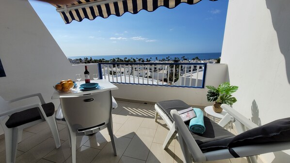A balcony with a table set for two, a chair, a small potted plant, and a view of the sea and buildings.