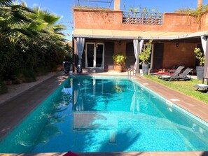 A rectangular swimming pool with clear blue water, surrounded by a tiled deck. There are lounge chairs and a potted plant near the pool, and a building with a balcony in the background. - Palais Akki (Marrakesh)