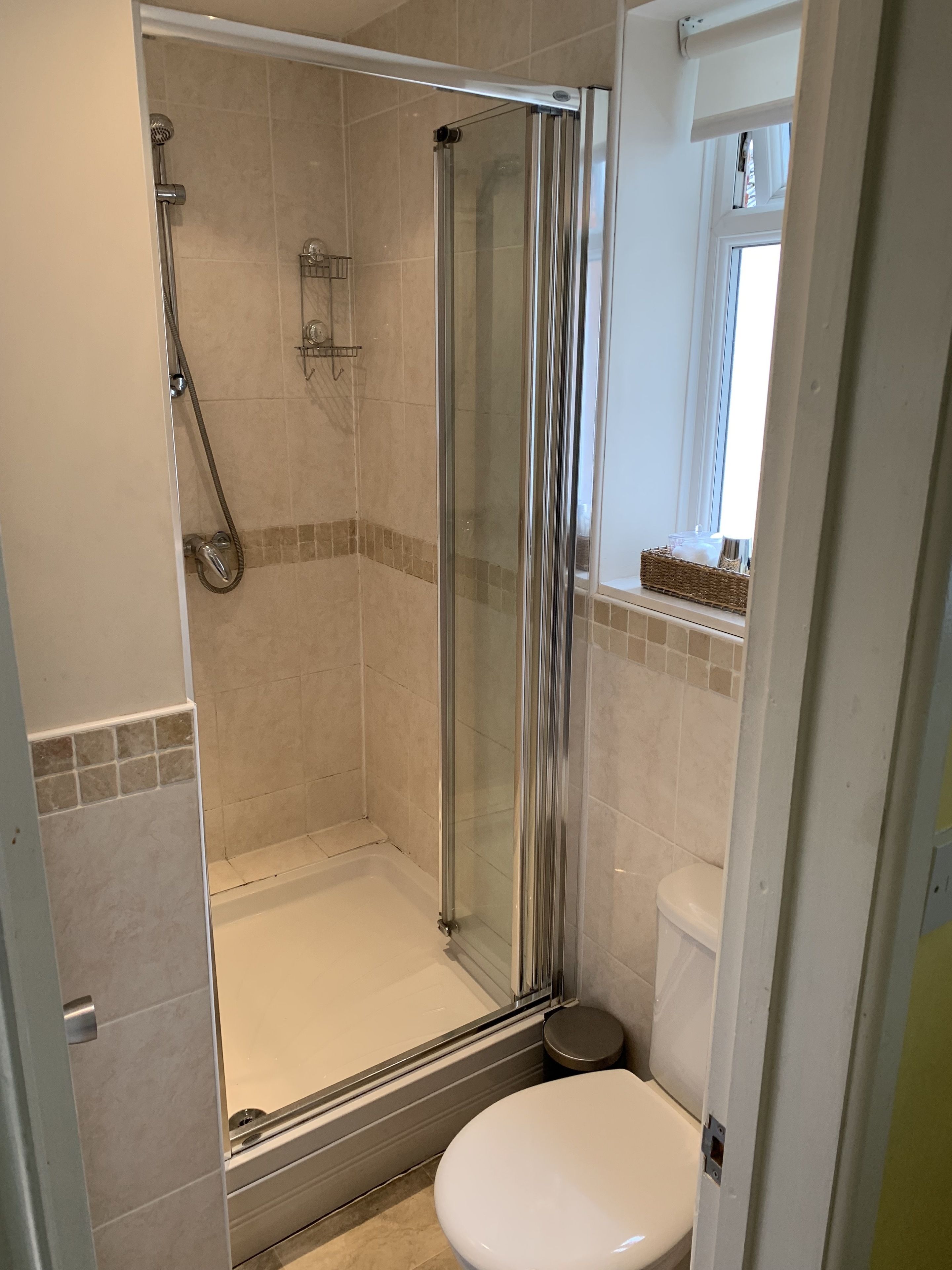 Eine Dusche mit Glastür, eine Toilette und ein kleines Fenster mit Korb auf der Fensterbank.