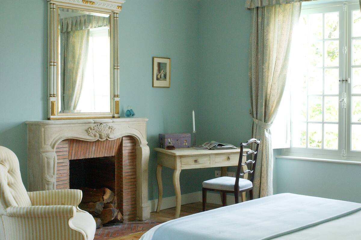 A bedroom with a bed, a chair, a desk, a fireplace, and a window with curtains.