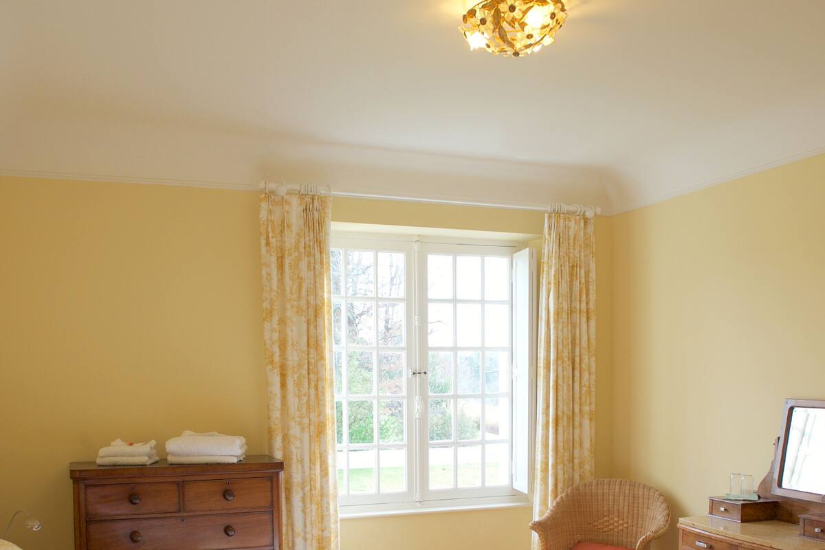 A bedroom with two beds, a wooden dresser, a wicker chair, a desk with a computer, and a window with curtains.