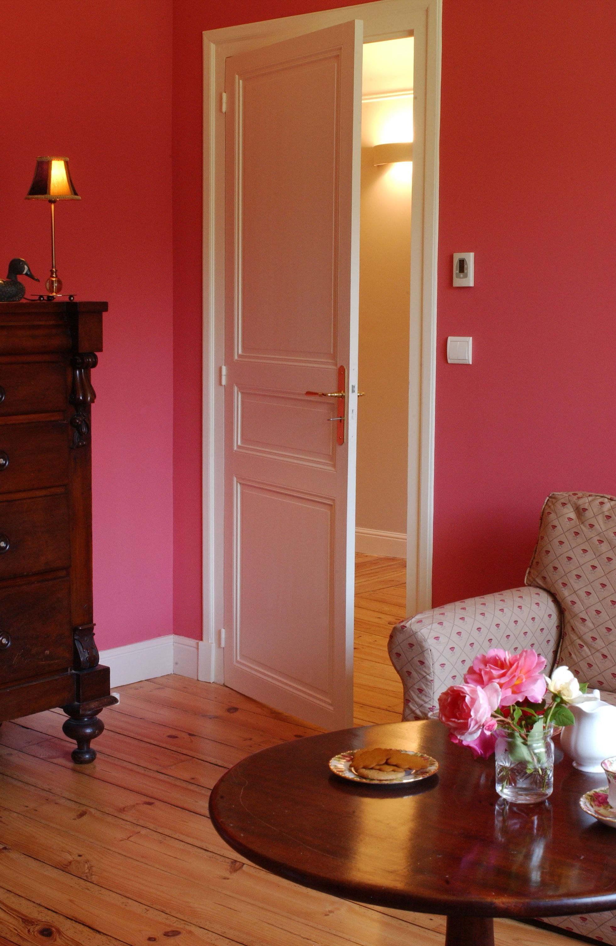 Una habitación con un aparador de madera, una mesa redonda con un plato y un jarrón de flores, una silla con estampado de corazones, y una puerta que da a otra habitación.
