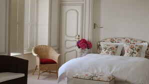 A bedroom with a bed, a wicker chair with a red cushion, a small table with a vase of flowers, and a door with a handle. - Clos Mirabel Manor B&B (Jurancon)