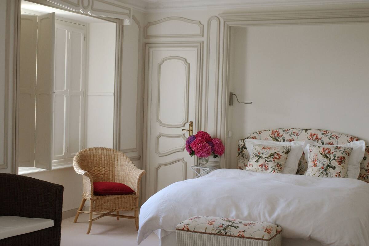 A bedroom with a bed, a wicker chair with a red cushion, a small table with a vase of flowers, and a door with a handle.