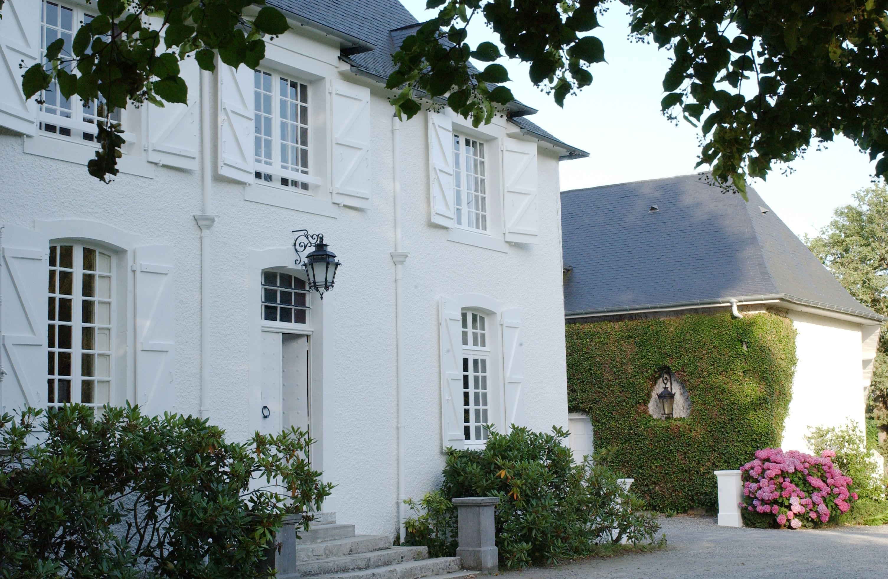 A white building with multiple windows and a door, surrounded by greenery and a large bush of pink flowers.