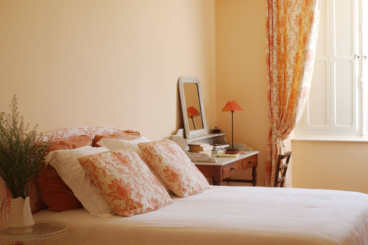 A neatly made bed with floral pillows, a bedside table with a lamp, a mirror, and a window with curtains.