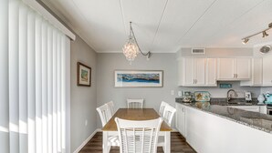 Un coin repas avec une table en bois et des chaises blanches, un lustre, et une cuisine dotée de meubles blancs et d’un plan de travail en granit.