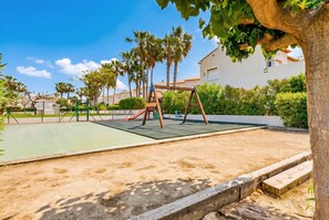 A playground with a slide and swings, surrounded by palm trees and a building in the background.