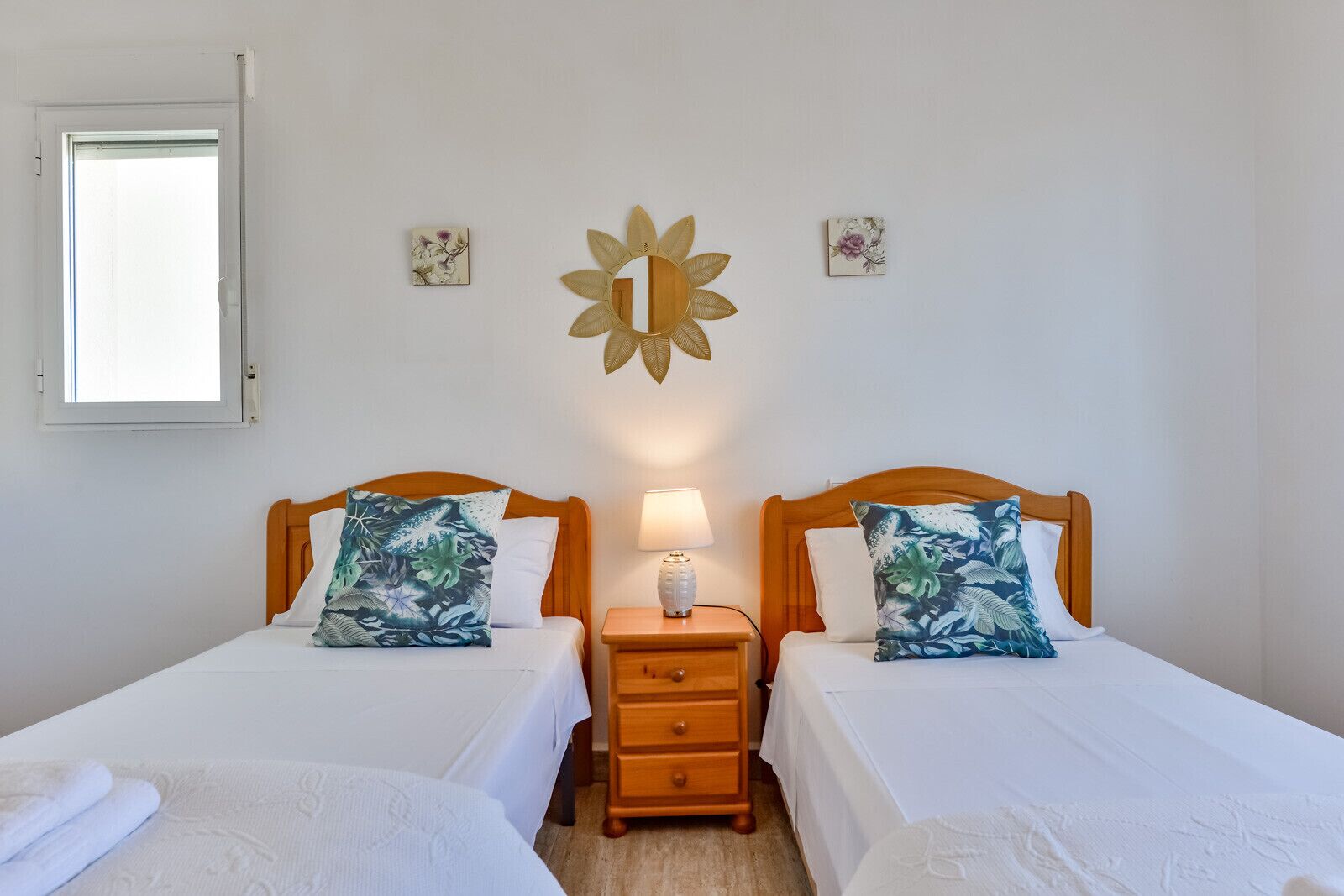 Ein Doppelzimmer mit zwei Einzelbetten, jeweils mit Holz-Kopfteil, weißer Bettwäsche und Kissen mit tropischem Blattmuster. Ein Nachttisch mit Lampe und ein dekorativer, sonnenförmiger Spiegel befinden sich zwischen den Betten.