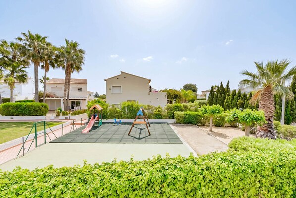 A playground with a slide and swings, surrounded by greenery and a building in the background. - Bungalow rental in Calpe, MARGARET (Calpe)