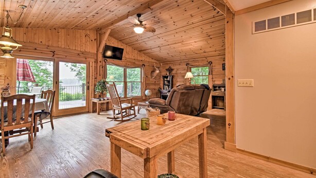 Interior de una cabaña de madera con comedor, sala de estar con sofá y chimenea, y un ventilador de techo.