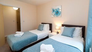 A hotel room with two beds, each with a blue blanket and white pillows, a bedside table with a lamp, and a painting on the wall. - Bladon Court (Basingstoke)