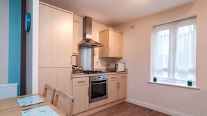 A compact kitchen with wooden cabinets, a gas stove, a microwave, and a window with curtains. - Bladon Court (Basingstoke)