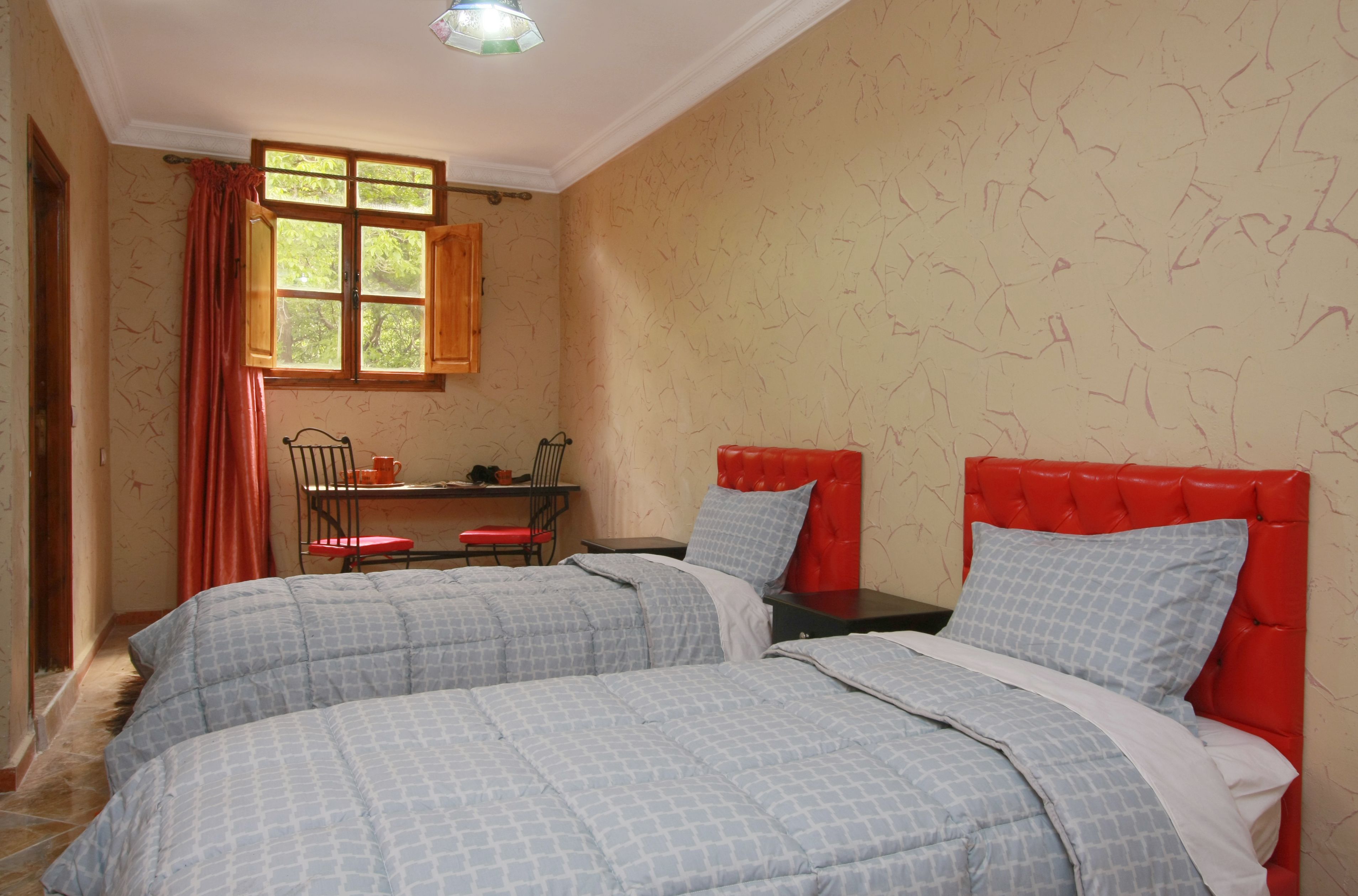 A bedroom with two beds, a window with curtains, a small table with a chair, and a red headboard.