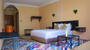 A hotel room with a bed, a small table, a colorful rug, a nightstand, and a wall-mounted shelf. - Kasbah Imoula (Asni)